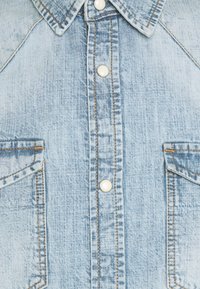 Hellblaues Denim-Hemd mit weißen Druckknöpfen, zwei Brusttaschen mit Klappen und orangefarbenen Nähten an den Säumen und dem Kragen.