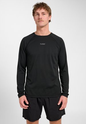 Mężczyzna z krótkimi włosami i wąsami, ubrany w czarną koszulkę z długim rękawem z logo "hummel" oraz czarne szorty, stoi na jednolitym jasnym tle.