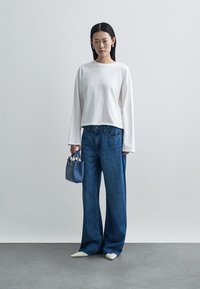 Weißes Langarmoberteil, blaue Jeans mit weitem Bein und eine kleine blaue Handtasche. Das Modell trägt spitze weiße Schuhe; minimalistisches Design und Farben.