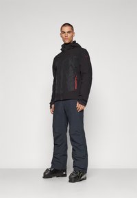 Schwarze isolierte Jacke mit einem strukturierten oberen Panel, roten Akzenten und einer Reißverschluss-Tasche, kombiniert mit dunkelblauen Hosen und schwarzen Skischuhen.
