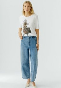 Witte graphic t-shirt met een bedrukt ontwerp, gecombineerd met lichtblauwe, wijde jeans met hoge taille. Model draagt naakte hakken.