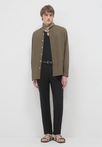 Olivgrünes Hemd mit Stehkragen, schwarzes T-Shirt, schwarze Hose und beiges Sandalen mit Riemen, ergänzt durch einen gemusterten Schal.