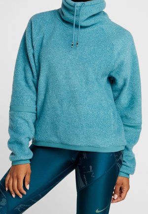 Person wearing a teal high-neck fleece pullover and dark blue patterned athletic leggings with a Nike logo, standing with hands relaxed.