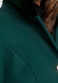 Cappotto verde in lana con superficie texturizzata, dotato di cappuccio, tasche laterali e chiusura con bottone dorato.