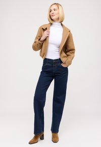 Giacca corta color tan, dolcevita bianca e jeans a gamba larga blu scuro. Stivaletti alla caviglia marroni con tacco. Tessuti lisci con vestibilità strutturata.