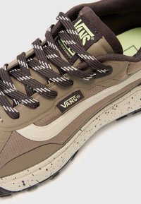 Zapatilla Vans en tonos bronceado y marrón, con tela texturizada, suela intermedia moteada y cordones con patrón. Logo exhibido de forma prominente en el lateral.