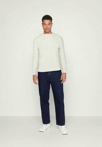 Weißer, gerippter Strickpullover, dunkelblaue Jeans und weiße Sneakers. Das Model steht mit entspannten Armen an den Seiten vor einem schlichten Hintergrund.