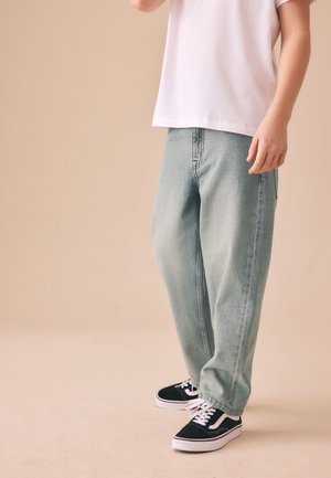 Person iført lyseblå løstsiddende jeans, sorte og hvide sneakers og en hvid skjorte, der står mod en beige baggrund.
