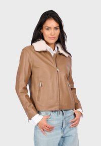 Vrouw in een tan leren jack met een wit schapenvachtkraag, witte shirtmanchetten en lichtblauwe high-waisted jeans, met de handen in de zakken.