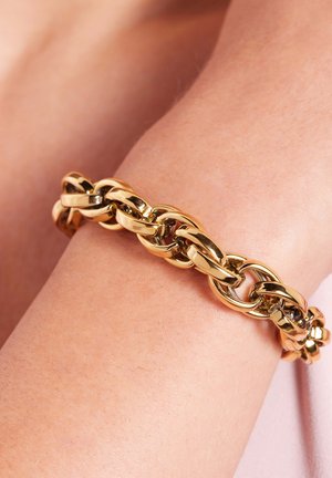 Gouden kettingarmband met in elkaar grijpende schakels, met een gladde, glanzende afwerking, gedragen om een pols tegen een lichte achtergrond.