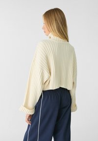 Pull en maille côtelée crème avec un col montant, un design court et des manches longues, associé à un pantalon bleu marine à taille élastique avec une passementerie blanche.