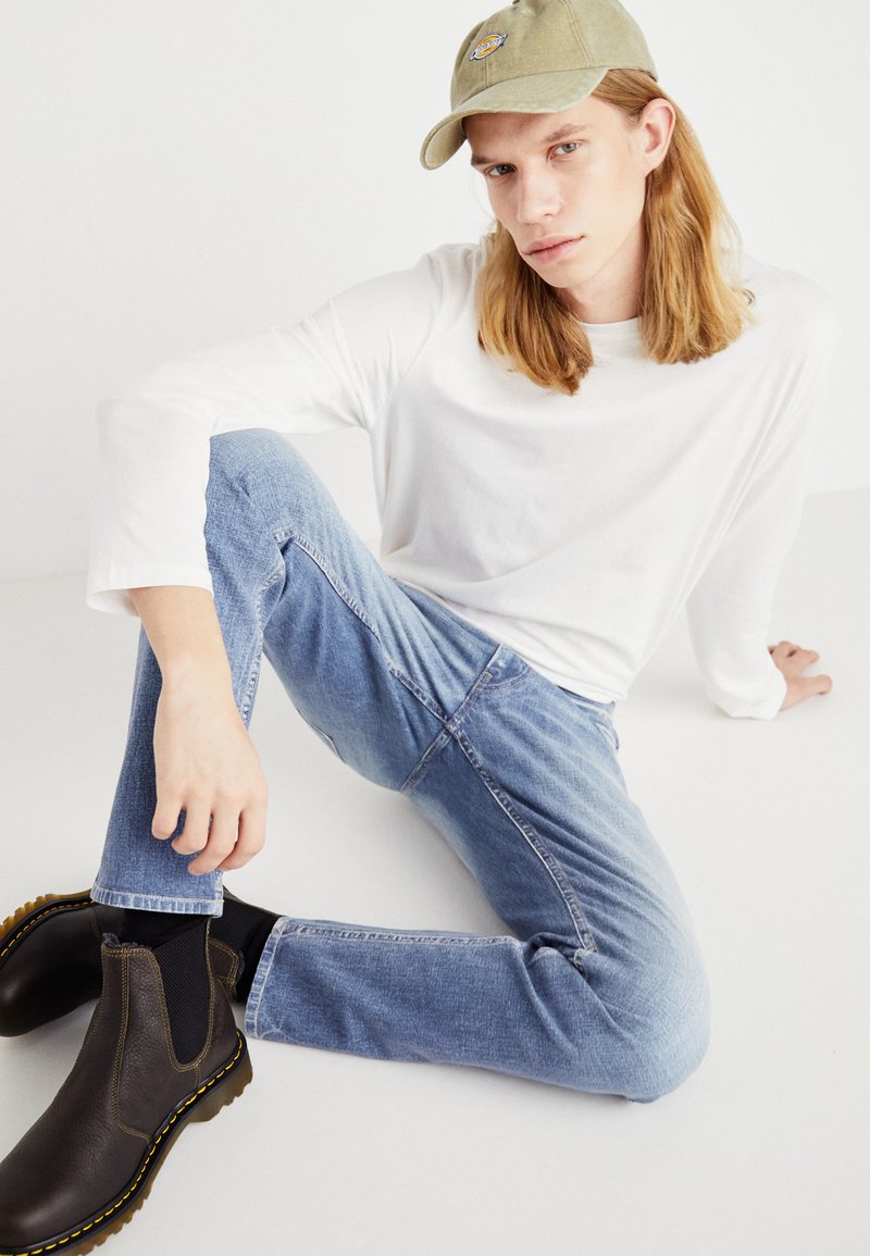 Leichtes, langärmeliges weißes Hemd; ausgebleichte blaue Jeans; dunkelbraune Chelsea-Boots; olivgrüne Mütze mit Logo; sitzende Pose vor einem weißen Hintergrund.