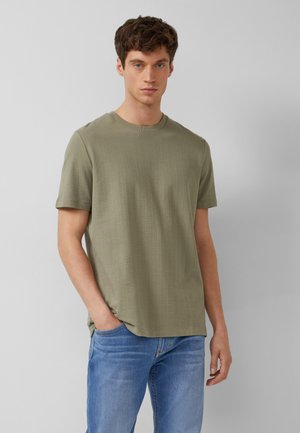 Młody mężczyzna z krótkimi brązowymi włosami, ubrany w oliwkową krótką koszulę i niebieskie dżinsy, stoi z jedną ręką w kieszeni.