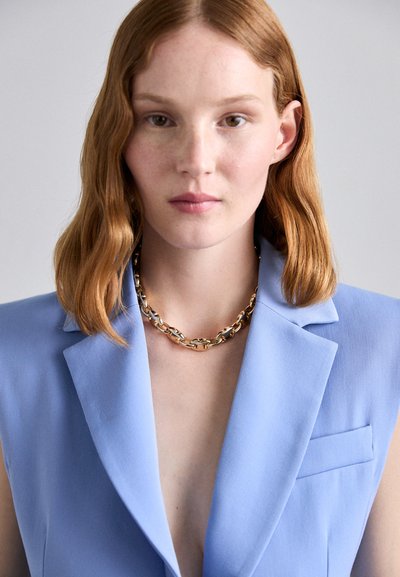 Light blue tailored blazer with a deep V-neck. Gold chain necklace accents the neckline. Smooth fabric with structured lapels.