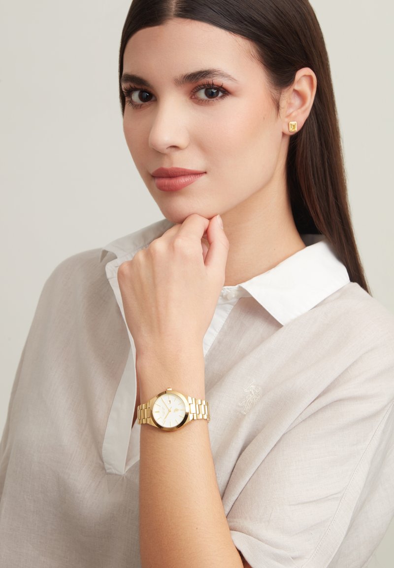 Montre-bracelet en or avec un cadran rond, un cadran blanc et un bracelet en maillons dorés, portée sur une main reposant près du menton. Chemise en tissu léger de couleur gris clair.