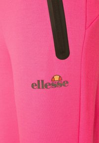 Roze sportbroek van gladde stof, met een zwarte ritssluiting en een grijs "ellesse" logo met een embleem.