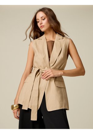 Mujer que lleva blazer beige sin mangas con cinturón en la cintura y pantalones negros, posando con la mano cerca de la cintura y una gran pulsera dorada en la muñeca.