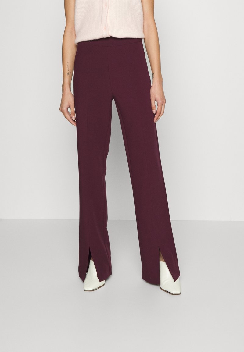 Femme portant un pantalon prune taille haute à jambes larges avec des fentes devant, associé à des bottes blanches à bouts pointus et un haut sans manches rose clair.