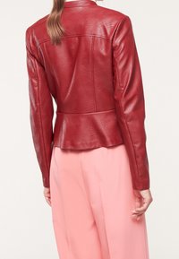 Kobieta w dopasowanej czerwonej skórzanej kurtce i luźnych różowych spodniach, pokazana z tyłu na jasnym tle.