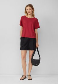T-shirt rosso a maniche corte abbinato a pantaloni neri, borsa a tracolla nera e sandali marroni. Il tessuto appare morbido, con un design casual e una vestibilità rilassata.