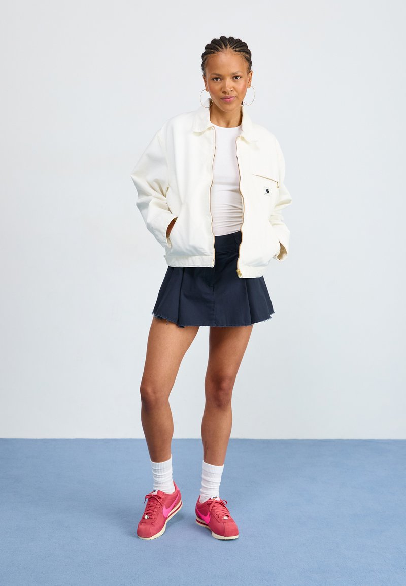 Veste blanche zippée, chemise blanche ajustée, jupe plissée bleu marine, baskets roses, chaussettes blanches. Le modèle se tient devant un fond clair.