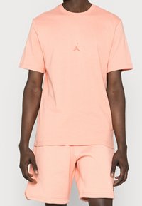 Camiseta de manga corta color durazno con cuello redondo, que presenta un sutil logo de Jordan en el pecho. Tejido suave y corte relajado apropiado para el uso diario.