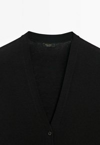 Schwarze Strickjacke mit V-Ausschnitt, verfügt über eine glatte Textur und einen einzelnen Knopfverschluss vorne. Das Etikett ist innen sichtbar.