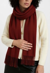Sciarpa bordeaux realizzata in morbido tessuto, con estremità frangiate, avvolta attorno al collo, abbinata a un cardigan color crema.