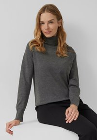 Femme aux cheveux châtain clair portant un pull à col roulé gris et un pantalon noir, assise sur un bloc blanc contre un fond gris clair.