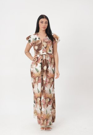 Vrouw staand met één hand op heup, draagt een witte jumpsuit met grote bruine en perzikkleurige bloemenprints en sandalen met open tenen.