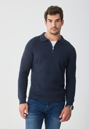Man wearing navy blue ribbed zip-up sweater, white undershirt, blue jeans, and brown wristwatch, standing against a plain light background.