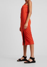 Hihaton oranssi mekko, jossa on nappi etuosa, polvimittainen leikkaus ja sivureikä. Valmistettu teksturoidusta kankaasta ja yhdistetty mustiin sandaaleihin.