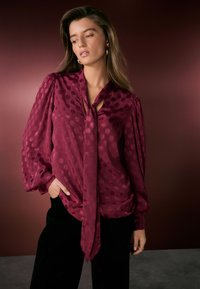 Blusa bordeaux con motivo a pois, dettaglio con fiocco al collo, maniche a sbuffo e vestibilità morbida. Abbinata a pantaloni neri su sfondo neutro.