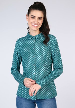 Green patterned shirt featuring a subtle geometric design, long sleeves, a collar, and button-down front. Paired with blue jeans.