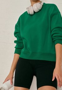 Groene katoen sweatshirt met geribbelde manchetten, gecombineerd met zwarte geribbelde fietsshorts. Witte koptelefoon rustend op de nek, met een waterfles in de hand.