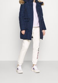 Giacca puffer blu con cappuccio foderato in pelliccia, design trapuntato, tasche frontali; abbinata a pantaloni della tuta color crema e sneaker bianche con accenti viola.