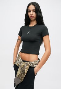 Λεπτή γυναίκα με μακριά μαύρα μαλλιά, φορώντας μια στενή μαύρη κοντή μπλούζα με ένα μικρό λευκό λογότυπο και ένα στρατιωτικό μπουφάν δεμένο γύρω από τη μέση της.