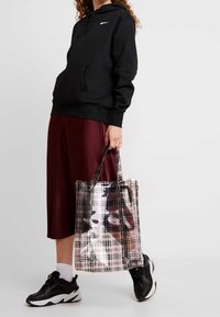 Person som bär en svart huvtröja, burgundisk kjol, svarta sneakers och håller en genomskinlig rutmönstrad tote-väska med synliga innehåll mot vit bakgrund.