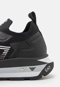 Svart sportskor med en texturerad stickad ovandel, gummisula och påfallande logointryck i guld och silver på hälen.