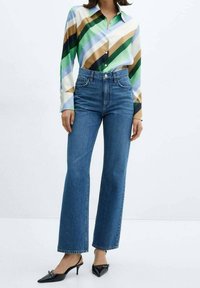Frau trägt hochtaillierte blaue Jeans, ein mehrfarbig gestreiftes Hemd mit diagonalen Streifen und schwarze spitze Slingback-Schuhe mit kitten heels.