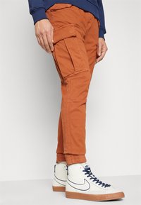 Noge v rjavorjavi cargo hlačah z velikimi žepi in belih supergah Nike visokih z modrimi detajli, stojijo na belem tleh.