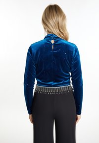 Blauwe fluwelen lange mouwen top met een sleutelgat aan de achterkant, voorzien van glinsterende accenten, gecombineerd met zwarte broeken versierd met zilveren kraalfranje.