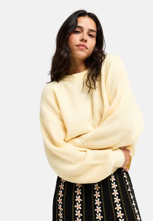 Junge Frau mit dunklen Haaren, die einen hellgelben, gerippten Pullover und einen schwarzen Rock mit vertikalen Blumenmustern trägt und mit verschränkten Armen posiert.