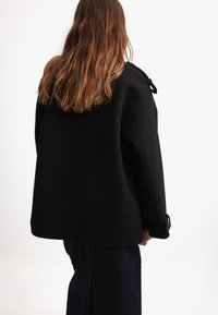 Cappotto in misto lana nero con vestibilità morbida, dotato di un ampio colletto e dettagli a bottoni. La parte posteriore presenta un design pulito e dettagli minimal.