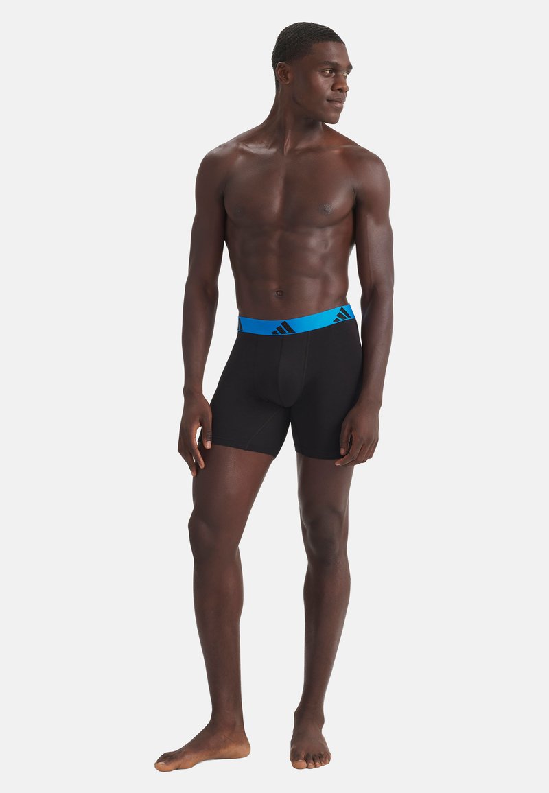Homme mince à la peau foncée debout pieds nus, portant un boxer noir avec une ceinture bleue, regardant vers la droite sur un fond blanc.