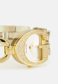 Gold-tone watch featuring a large G-shaped case adorned with rhinestones, a white dial, and a chunky linked bracelet design.