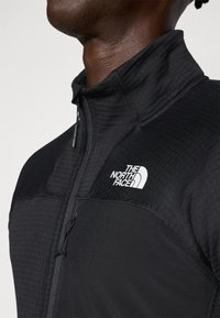 Svart zip-up jakke med teksturert stoff, glatte detaljer og hvit logo på brystet. Designet for varme og komfort.