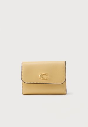 Kompakt beige læderpung med afrundet klap og guldtonet "C" emblem placeret i midten på forsiden mod ensfarvet baggrund.