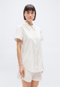 Femme aux cheveux courts et foncés, portant une chemise blanche à manches courtes boutonnée et un short assorti avec un ourlet festonné jaune, debout devant un fond uni.