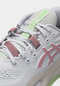 Άσπρο αθλητικό παπούτσι Asics με ροζ λεπτομέρειες, άσπρα κορδόνια, αναπνεύσιμη επάνω επιφάνεια από δίχτυ και ετικέτα γλώσσας με λογότυπο σε ανοιχτό πράσινο χρώμα.